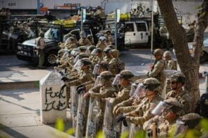 California National Guard soldiers in camouflage uniforms and full riot gear, including helmets and face shields, form a line on a city street in Los Angeles. They are holding batons and large, clear riot shields.