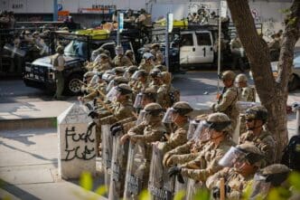 California National Guard soldiers in camouflage uniforms and full riot gear, including helmets and face shields, form a line on a city street in Los Angeles. They are holding batons and large, clear riot shields.