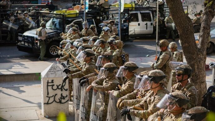 California National Guard soldiers in camouflage uniforms and full riot gear, including helmets and face shields, form a line on a city street in Los Angeles. They are holding batons and large, clear riot shields.