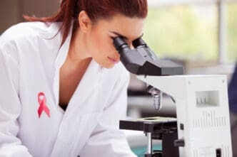 A female scientist with red hair, wearing a white lab coat with a pink breast cancer awareness ribbon pin, looks intently through a microscope in a laboratory setting.