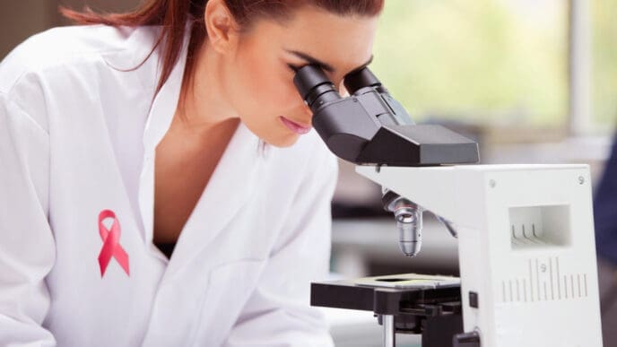 A female scientist with red hair, wearing a white lab coat with a pink breast cancer awareness ribbon pin, looks intently through a microscope in a laboratory setting.