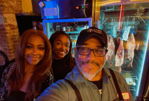 A selfie of Chef Russell Jackson, wearing a cap and glasses, with two female companions. They are smiling in what appears to be a restaurant kitchen, with a commercial dry-aging cabinet containing hanging poultry visible in the background.