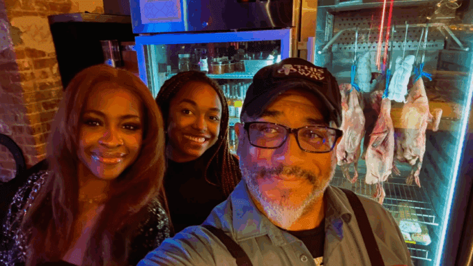 A selfie of Chef Russell Jackson, wearing a cap and glasses, with two female companions. They are smiling in what appears to be a restaurant kitchen, with a commercial dry-aging cabinet containing hanging poultry visible in the background.