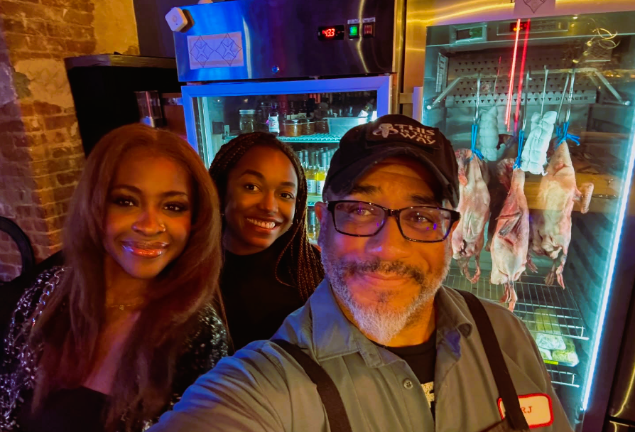 A selfie of Chef Russell Jackson, wearing a cap and glasses, with two female companions. They are smiling in what appears to be a restaurant kitchen, with a commercial dry-aging cabinet containing hanging poultry visible in the background.