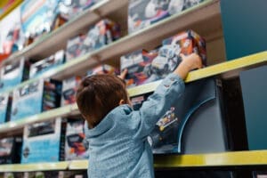 A young boy, seen from behind, stretches upwards with both hands to reach a toy box on a high shelf in a brightly lit toy store aisle.