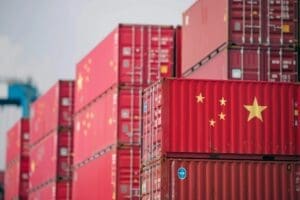 A close-up photo of a red shipping container marked with the flag of China, stacked among many other containers at a busy shipping port.