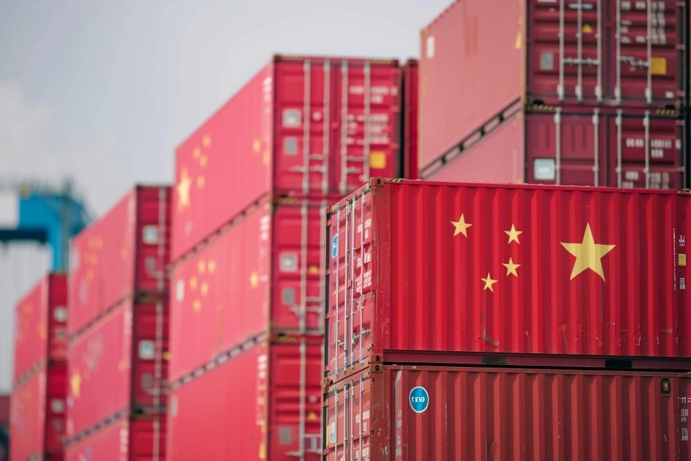 A close-up photo of a red shipping container marked with the flag of China, stacked among many other containers at a busy shipping port.