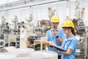 Two industrial workers, a man and a woman who appear to be of East Asian descent, wearing yellow hard hats and blue uniforms, inspect a clipboard and talk in a factory setting with machinery in the background.