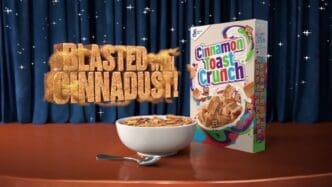A commercial product shot of Cinnamon Toast Crunch cereal, featuring a full bowl of cereal and the product box on a stage. A large 3D text graphic reads, "BLASTED with CINNADUST!" against a sparkly blue curtain background.