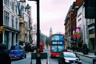 A bustling London street scene featuring a red double-decker bus in the foreground and the iconic Big Ben clock tower visible in the distance. Historic buildings line both sides of the street under an overcast sky.