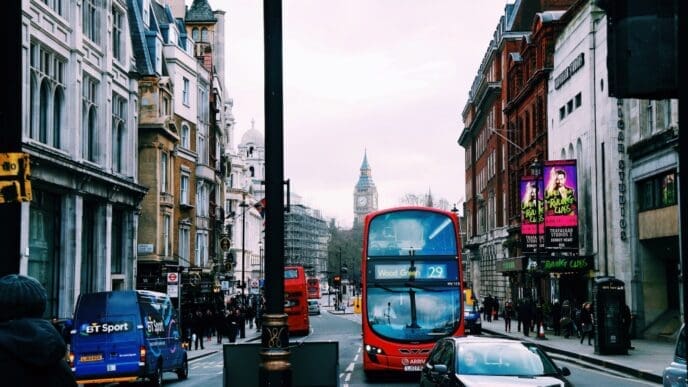 A bustling London street scene featuring a red double-decker bus in the foreground and the iconic Big Ben clock tower visible in the distance. Historic buildings line both sides of the street under an overcast sky.