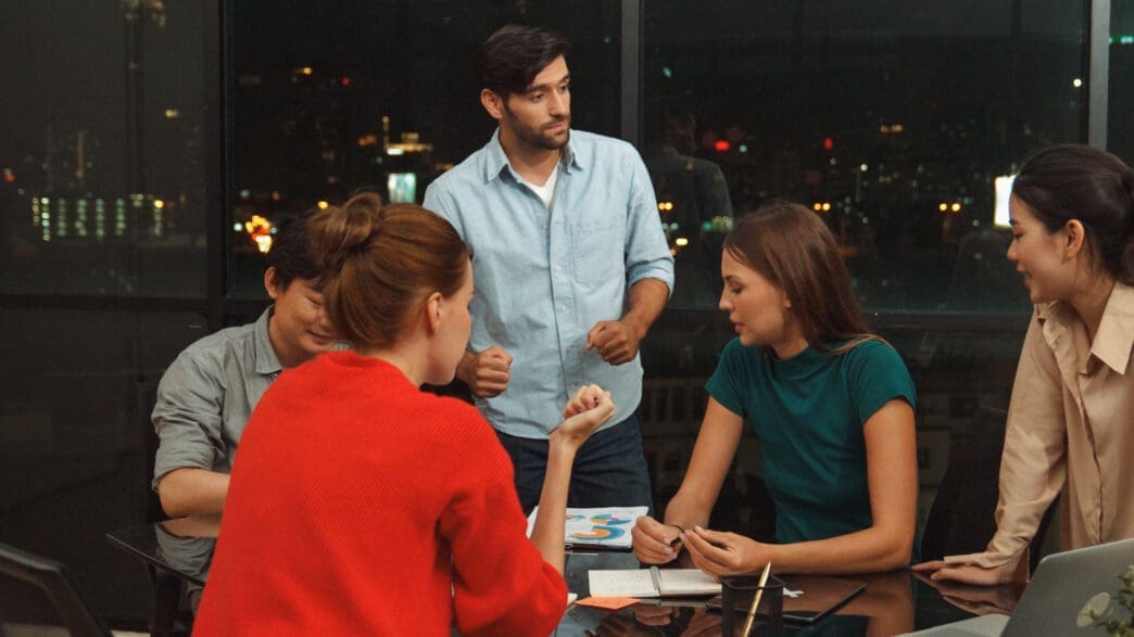 A diverse group of five people are gathered around a table in a modern office at night, engaged in an active discussion, with city lights visible outside the windows.