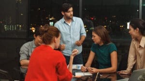 A diverse group of five people are gathered around a table in a modern office at night, engaged in an active discussion, with city lights visible outside the windows.