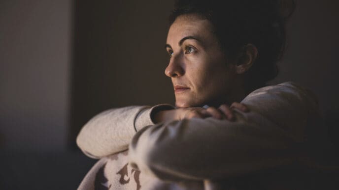 A close-up portrait of a woman with a serious, contemplative expression, looking into the distance with her arms crossed, in a dimly lit setting.