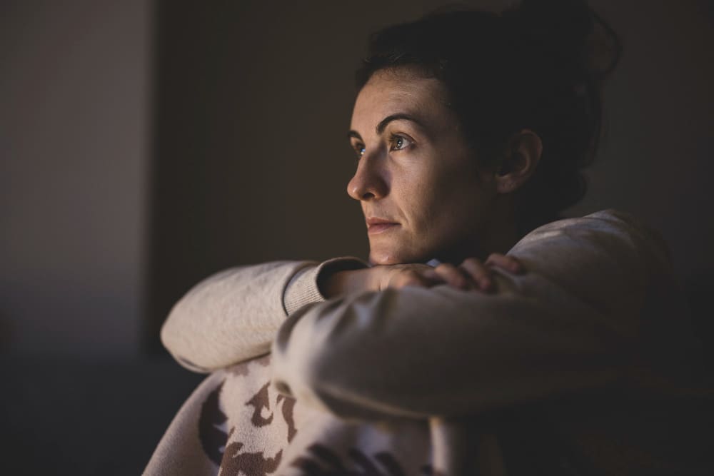 A close-up portrait of a woman with a serious, contemplative expression, looking into the distance with her arms crossed, in a dimly lit setting.