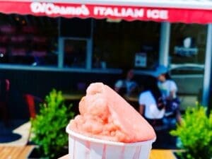 A person's hand holds a paper cup of pink Italian ice in the foreground, with the sunny, out-of-focus storefront of DiCosmo's Italian Ice visible in the background.