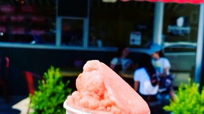 A person's hand holds a paper cup of pink Italian ice in the foreground, with the sunny, out-of-focus storefront of DiCosmo's Italian Ice visible in the background.