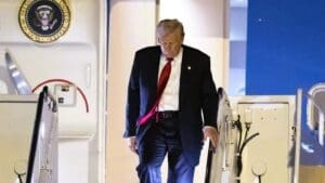 President Donald Trump, wearing a suit and red tie, walks down the stairs of Air Force One at night, with the Presidential Seal visible on the plane's fuselage.