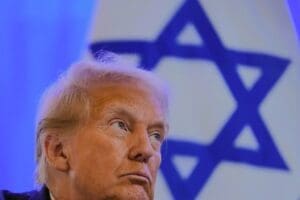 A close-up photograph of President Donald Trump with a serious expression, looking off-camera. The flag of Israel is visible and out of focus in the background.