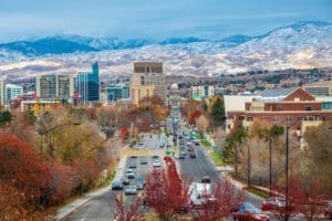 An elevated view of downtown Boise, Idaho, looking down a wide boulevard lined with autumn-colored trees towards the State Capitol Building and a skyline of modern buildings, with snow-capped mountains in the background.