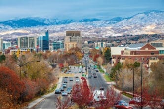 An elevated view of downtown Boise, Idaho, looking down a wide boulevard lined with autumn-colored trees towards the State Capitol Building and a skyline of modern buildings, with snow-capped mountains in the background.