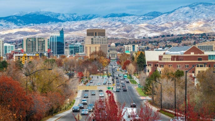 An elevated view of downtown Boise, Idaho, looking down a wide boulevard lined with autumn-colored trees towards the State Capitol Building and a skyline of modern buildings, with snow-capped mountains in the background.