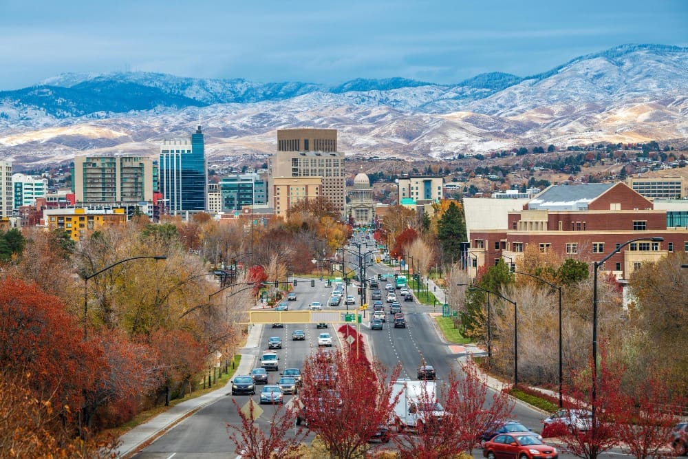 An elevated view of downtown Boise, Idaho, looking down a wide boulevard lined with autumn-colored trees towards the State Capitol Building and a skyline of modern buildings, with snow-capped mountains in the background.