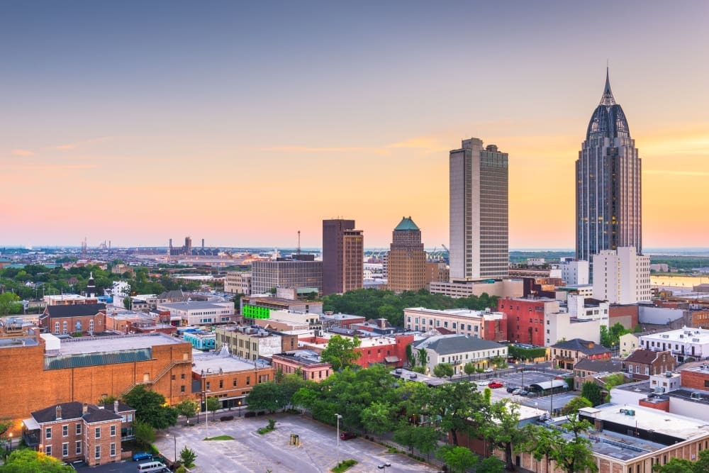An elevated view of the downtown skyline of Mobile, Alabama, at dusk or dawn, featuring prominent skyscrapers like the RSA Battle House Tower, a mix of historic and modern buildings, and a distant port area.