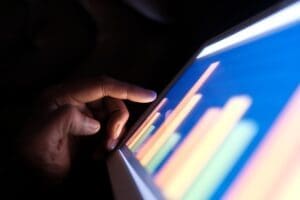 A close-up of a hand with a finger touching a glowing digital tablet screen that displays a colorful bar chart, symbolizing data analysis in a dark environment.