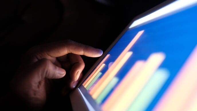 A close-up of a hand with a finger touching a glowing digital tablet screen that displays a colorful bar chart, symbolizing data analysis in a dark environment.