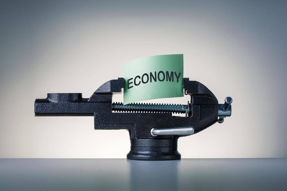 A conceptual image of a black bench vise squeezing a piece of green paper with the word "ECONOMY" printed on it, symbolizing economic pressure.