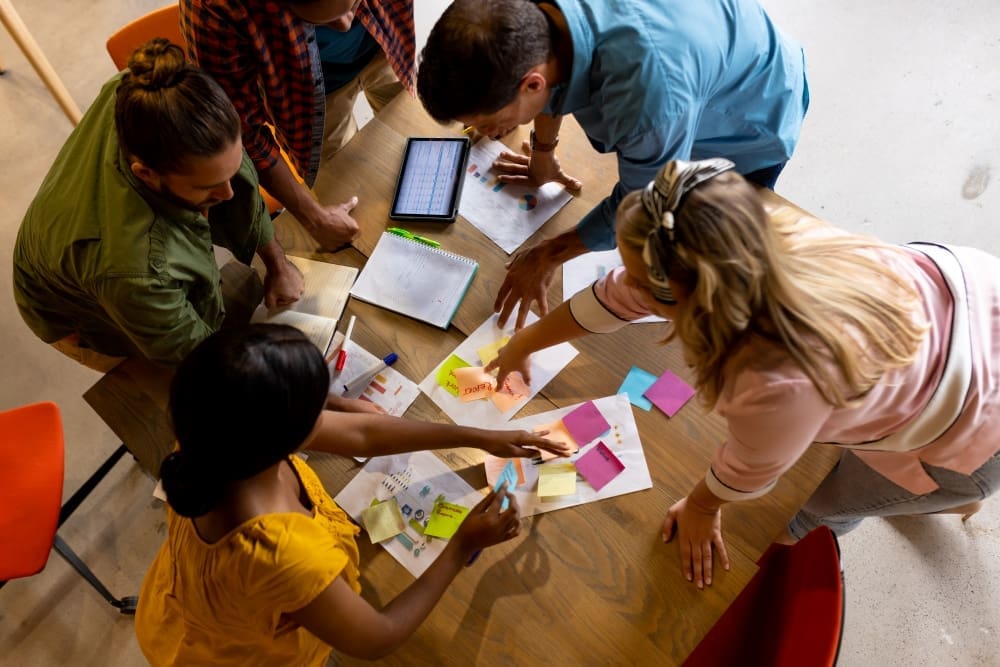 A high-angle view of a diverse group of educators and colleagues brainstorming around a wooden table covered in colorful sticky notes, papers, and a digital tablet.