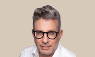 A professional headshot of Eldad Tamir, a middle-aged man with graying hair and black-framed glasses, wearing a white collared shirt and looking directly at the camera against a solid beige background.