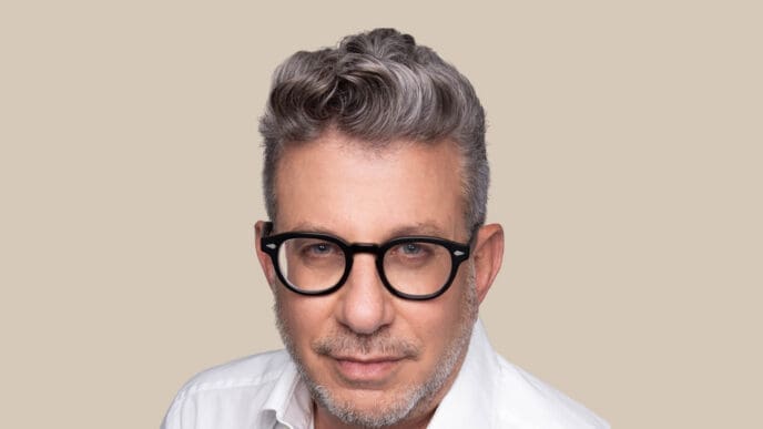 A professional headshot of Eldad Tamir, a middle-aged man with graying hair and black-framed glasses, wearing a white collared shirt and looking directly at the camera against a solid beige background.