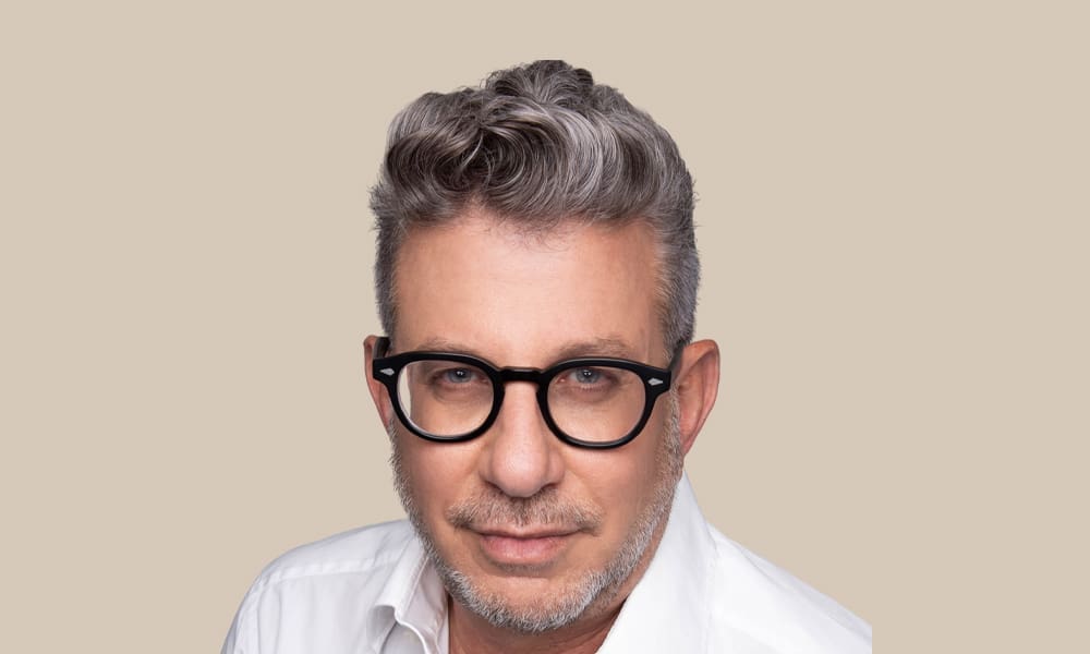 A professional headshot of Eldad Tamir, a middle-aged man with graying hair and black-framed glasses, wearing a white collared shirt and looking directly at the camera against a solid beige background.
