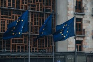 A low-angle view of the flags of European Union member states flying on flagpoles in front of the modern facade of the European Parliament building in Brussels, Belgium.
