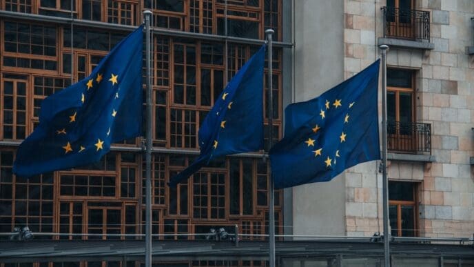 A low-angle view of the flags of European Union member states flying on flagpoles in front of the modern facade of the European Parliament building in Brussels, Belgium.