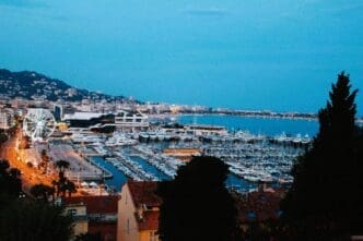 A high-angle photograph of the harbor in Cannes, France, taken at dusk. The image shows the harbor filled with yachts, a large illuminated Ferris wheel, and the lights of the city stretching along the coast.