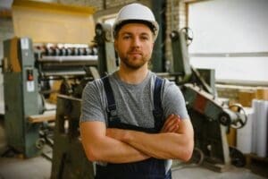 A man who appears to be White, wearing a white hard hat and blue overalls over a grey t-shirt, stands with his arms crossed in a factory setting with blurred machinery in the background.