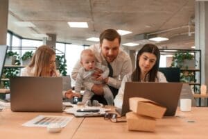 A father, is holding his infant son while standing between two female colleagues in a modern office. All three adults are collaborating and looking at a laptop screen together.
