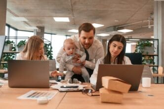 A father, is holding his infant son while standing between two female colleagues in a modern office. All three adults are collaborating and looking at a laptop screen together.