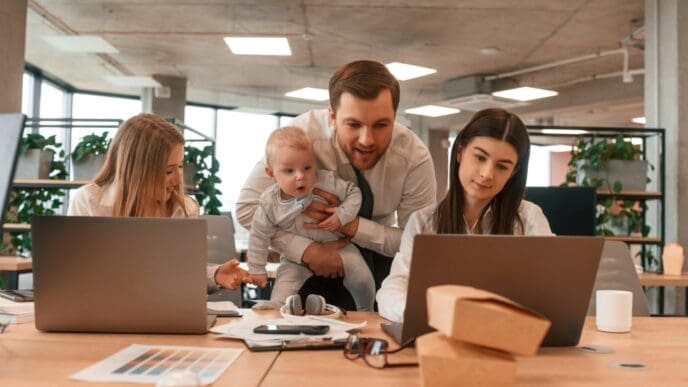 A father, is holding his infant son while standing between two female colleagues in a modern office. All three adults are collaborating and looking at a laptop screen together.