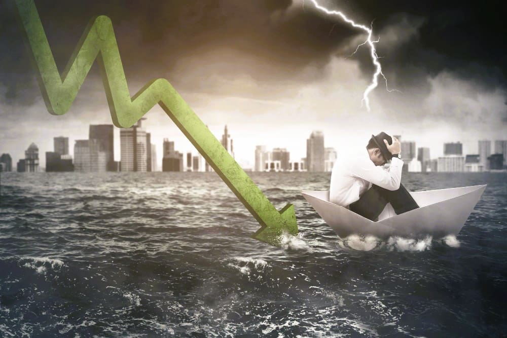 A conceptual image of a financial crisis, showing a man huddled in despair in a paper boat on turbulent waters under a stormy sky with lightning, while a large green graph arrow crashes into the water near a city skyline.