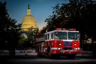 A bright red fire truck with illuminated emergency lights is parked in the foreground, with the iconic illuminated dome of the U.S. Capitol Building visible in the background at dusk, framed by dark trees.