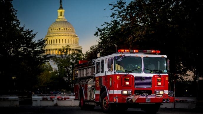 A bright red fire truck with illuminated emergency lights is parked in the foreground, with the iconic illuminated dome of the U.S. Capitol Building visible in the background at dusk, framed by dark trees.