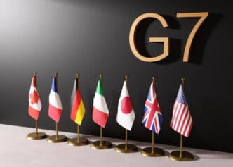 A 3D rendering of the national flags of the G7 countries—Canada, France, Germany, Italy, Japan, the UK, and the USA—standing together in a row, representing the international political forum.