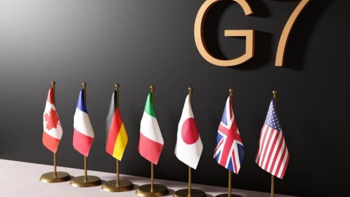 A 3D rendering of the national flags of the G7 countries—Canada, France, Germany, Italy, Japan, the UK, and the USA—standing together in a row, representing the international political forum.