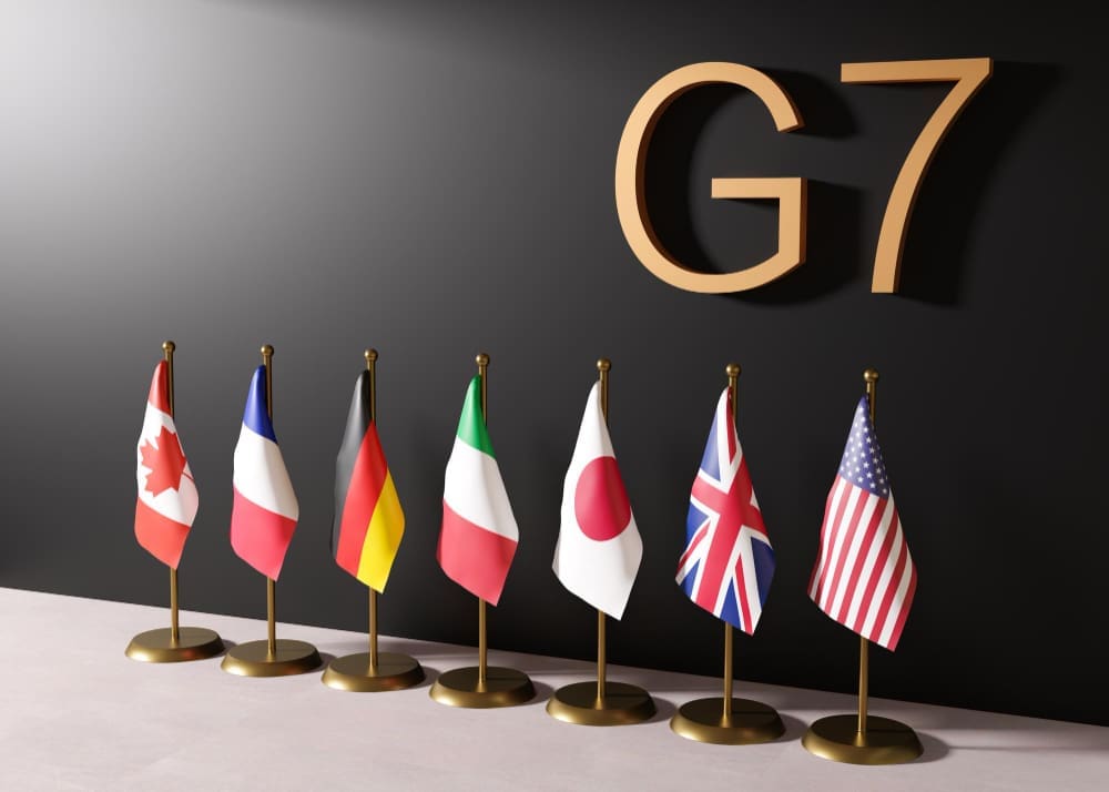 A 3D rendering of the national flags of the G7 countries—Canada, France, Germany, Italy, Japan, the UK, and the USA—standing together in a row, representing the international political forum.