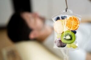A clear IV drip bag hangs from a stand, filled with colorful slices of various fruits like oranges, kiwi, blueberries, apples, and grapes, with a blurry figure of a person lying down in the background.