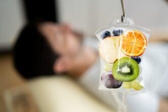A clear IV drip bag hangs from a stand, filled with colorful slices of various fruits like oranges, kiwi, blueberries, apples, and grapes, with a blurry figure of a person lying down in the background.
